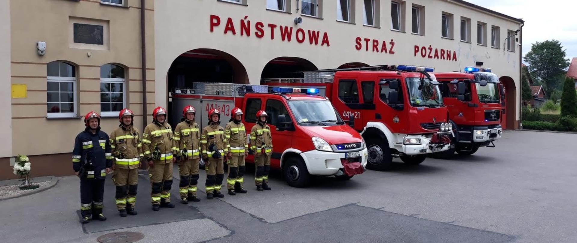 Strażacy ustawieni w szeregu i pojazdy pożarnicze z załączoną sygnalizacją przed budynkiem komendy.