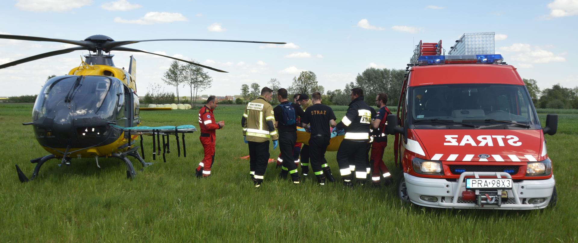 Łąka w Dębnie Polskim. Strażacy wraz z załogą śmigłowca LPR przenoszą chorego z wozu strażackiego, którym pacjent został przetransportowany z miejsca zamieszkania, do śmigłowca. Ze śmigłowca są wysunięte na zewnątrz nosze. W tle widoczne drzewa.