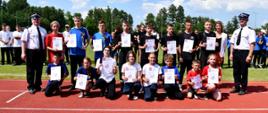 Na pierwszym planie dzieci z dyplomami za udział w zawodach po bokach osoby wręczające dyplomy, stoją na bieżni stadionu w tle na murawie pozostali zawodnicy zawodów.