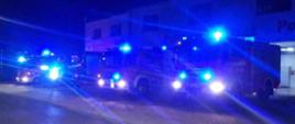 W hołdzie zmarłemu strażakowi. Zdjęcie przedstawia plac manewrowy przed budynkiem komendy. Na placu stoją 4 pojazdy pożarnicze z włączoną sygnalizacją świetlną. Jest pora nocna.