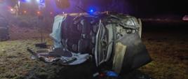 Uszkodzone auto leży na boku na łące w pobliżu drogi. W aucie odcięto dach.