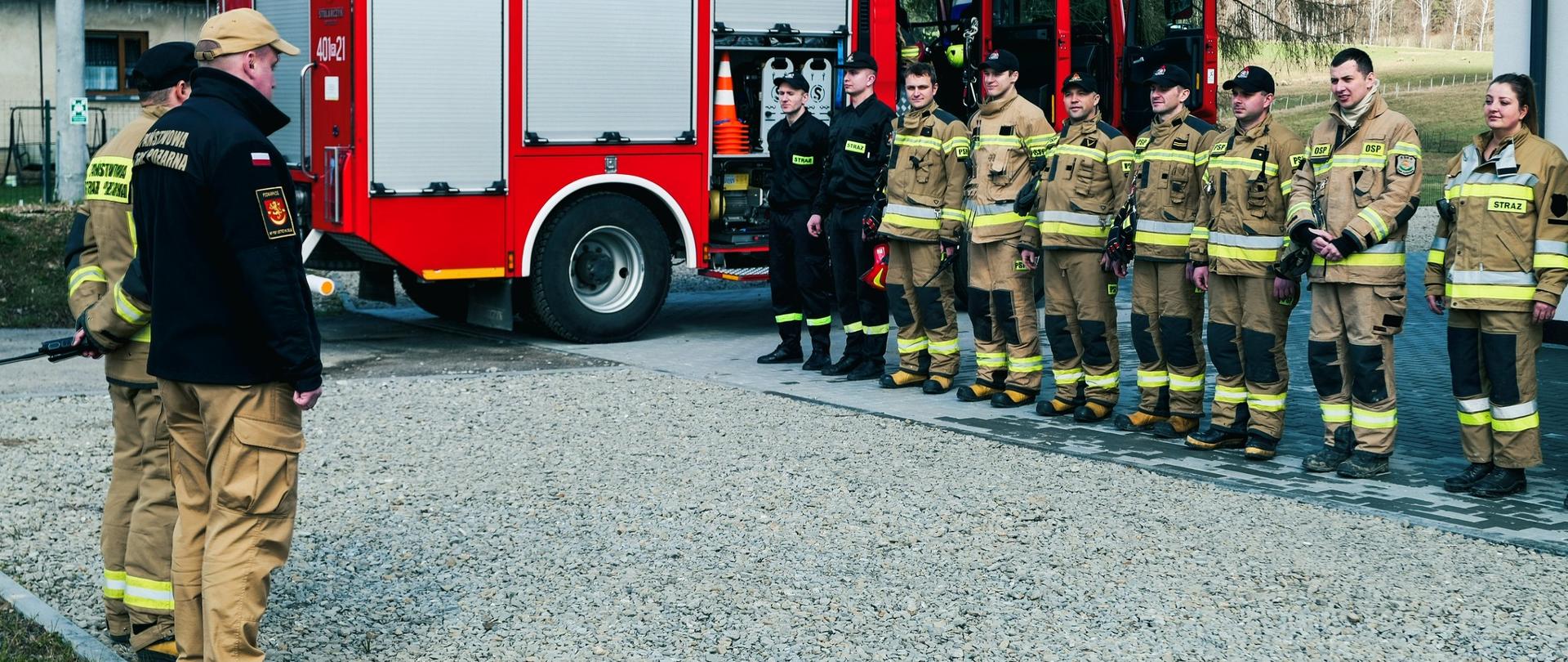 Zdjęcie przedstawia dowódców PSP stojących na przeciw strażaków ustawionych w szyku. W tle samochód pożarniczy