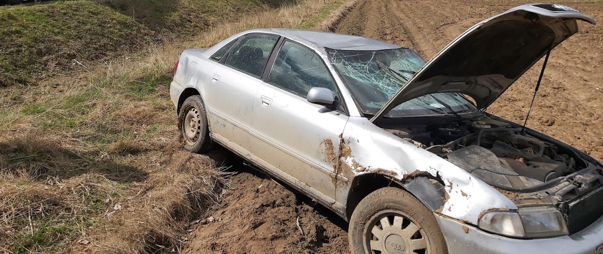 Zdjęcie przedstawia srebrny samochód osobowy z podniesioną maską i rozbitą szybką czołową, w tle widać karetkę pogotowia i samochód straży pożarnej 