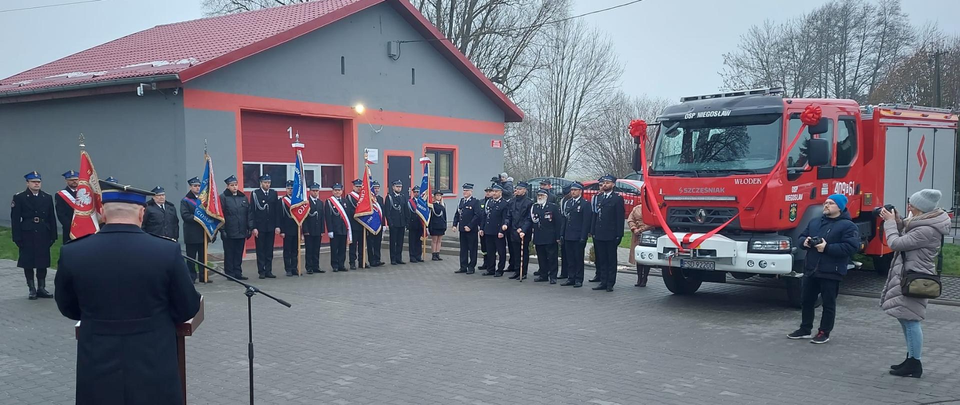 Fotografia przedstawia ujęcie placu apelowego z ustawionymi pocztami sztandarowymi oraz pododdziałem strażaków ochotników obok nowego samochodu gaśniczego.