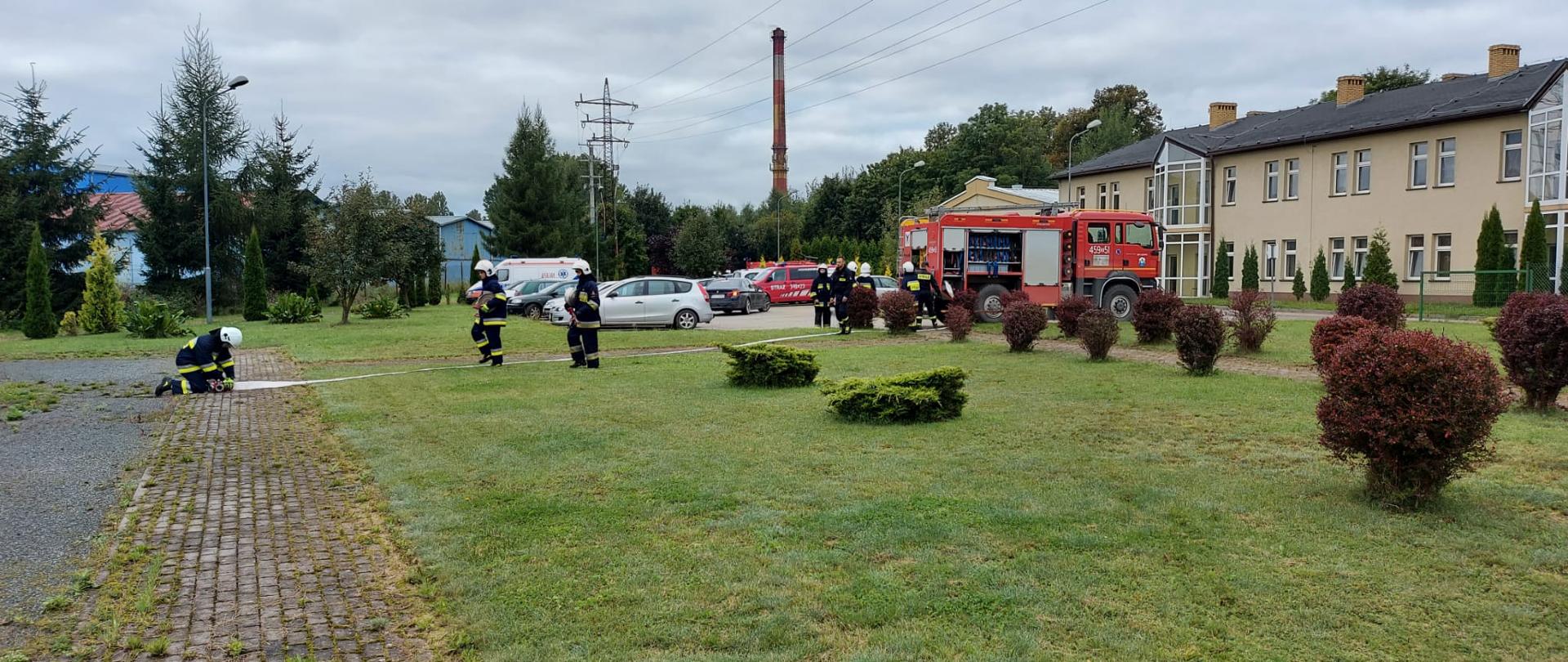 Strażacy Ochotniczej Straży Pożarnej podczas ćwiczeń na terenach otwartych. Po prawej stronie samochód ratowniczo gaśniczy o widocznym oznaczeniu 459-5. Druhowie rozstawiają linie gaśniczą zakończoną rozdzielaczem. Jeden ze strażaków podłącza odcinek do rozdzielacza . Dwóch idzie w jego kierunku niosąc węże gaśnicze. W tle dostrzegamy parking na którym stoją samochody osobowe.