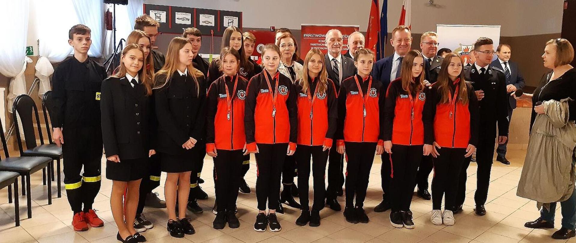 Uczestnicy uroczystości wręczenia promes dla młodzieżowych drużyn pożarniczych pozują do zdjęcia zbiorowego, wśród nich dzieci z młodzieżowej drużyny pożarniczej, strażacy druhowie, posłowie i senatorowie RP, przedstawiciele władz samorządowych.