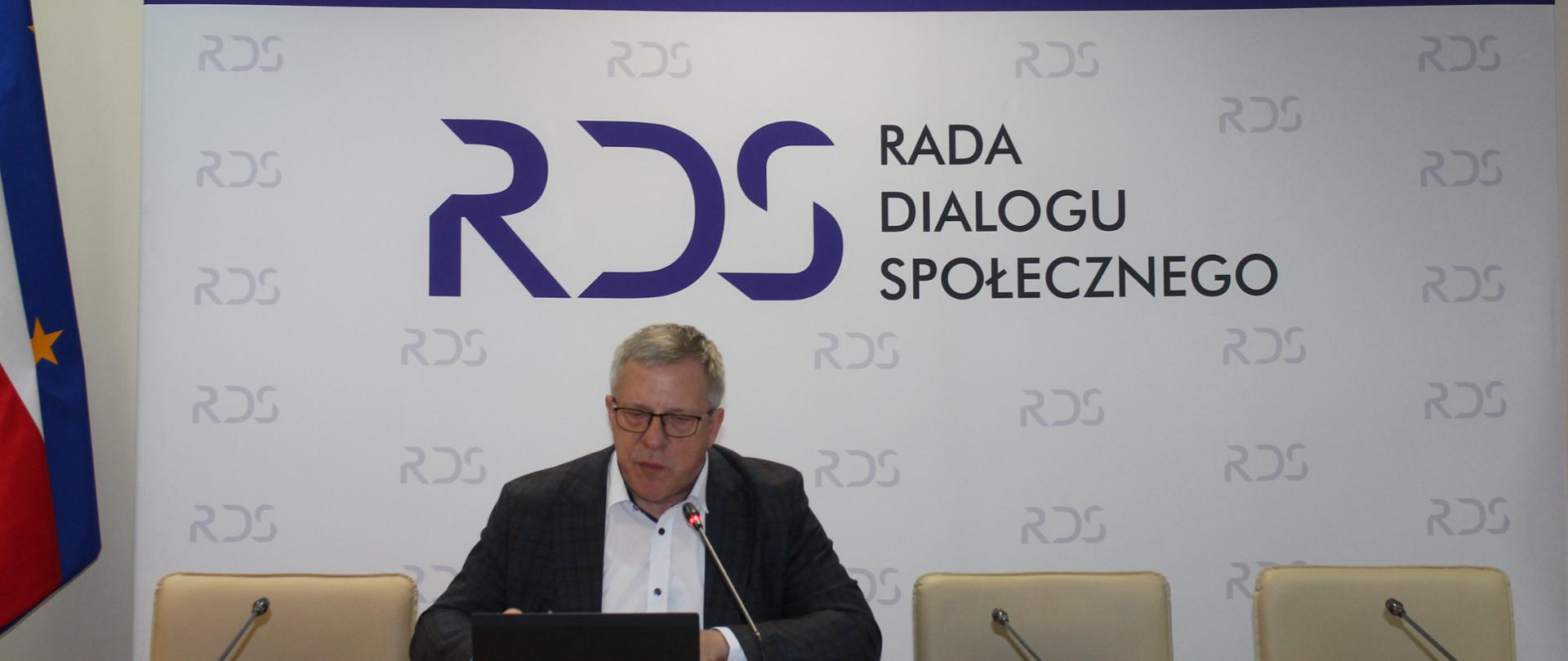 Zdjęcie przedstawia stół prezydialny za którym siedzi Pan Leszek Miętek, Przewodniczący Zespołu. W tle znajduje się baner z napisem Rada Dialogu Społecznego. Po lewej stronie stoją dwie flagi: Polski i UE. Nad banerem wisi godło Polski.