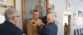 Strażacy Państwowej Straży Pożarnej stoją obok siebie strażak w mundurze oliwkowym otrzymuje od osoby cywilnej wyróżnienie.