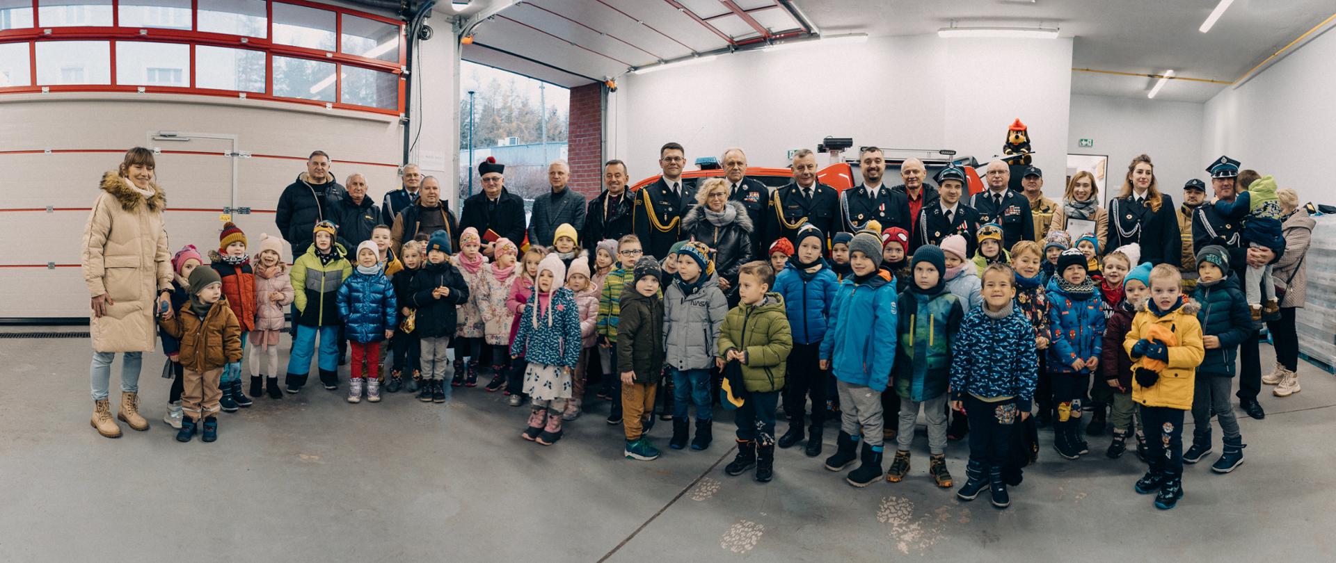 Zdjęcie grupowe funkcjonariuszy Państwowej Straży Pożarnej, druhów Ochotniczej Straży Pożarnej Duszniki-Zdrój oraz dzieci w garażu remizy strażackiej.