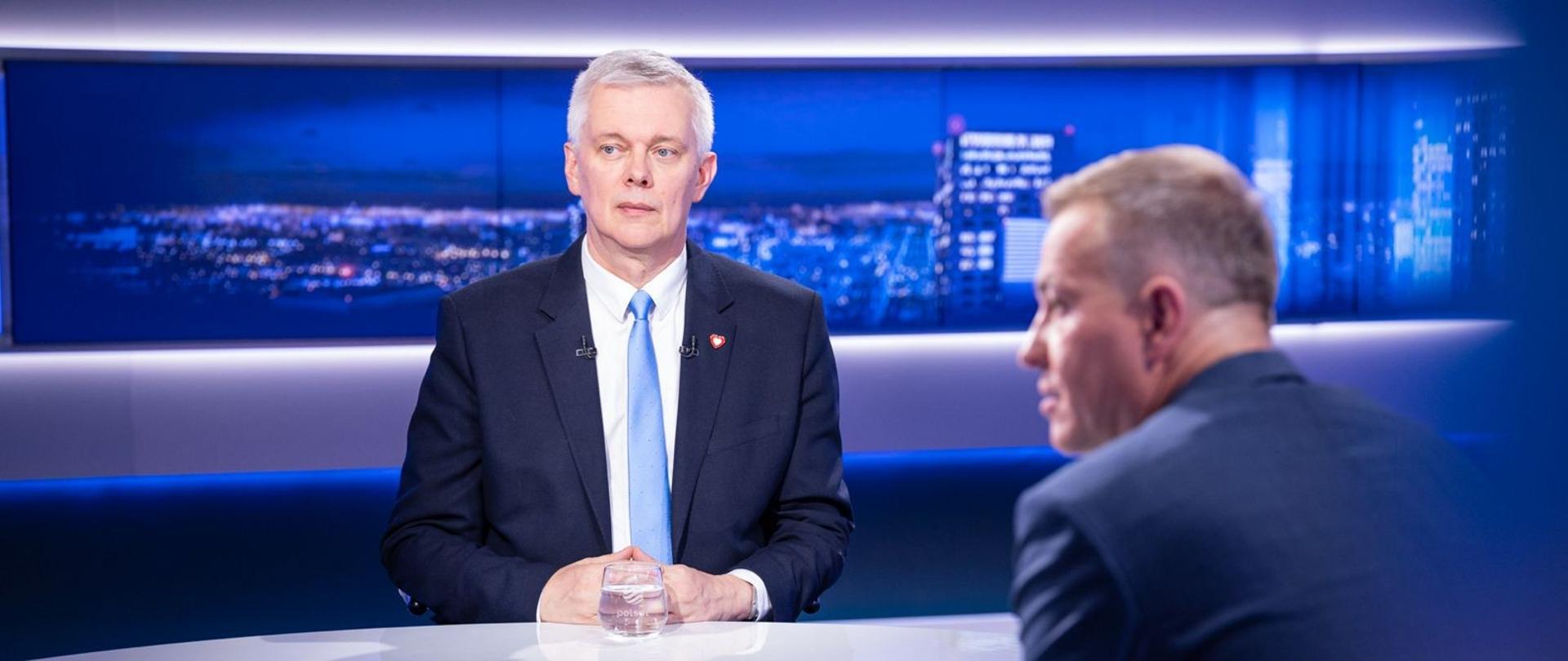 Mężczyzna w garniturze i jasnoniebieskim krawacie siedzi przy stole w studiu telewizyjnym. Naprzeciwko niego, częściowo tyłem do obiektywu, siedzi drugi mężczyzna. W tle widoczna jest niebieska panorama miasta.