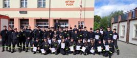 Na zdjęciu grupa 38 strażaków z ochotniczych straży pożarnych. Ubrani w czarne mundury koszarowe w ręku trzymają dyplomy ukończenia kursu. W tle budynek komendy powiatowej straży w Pucku.