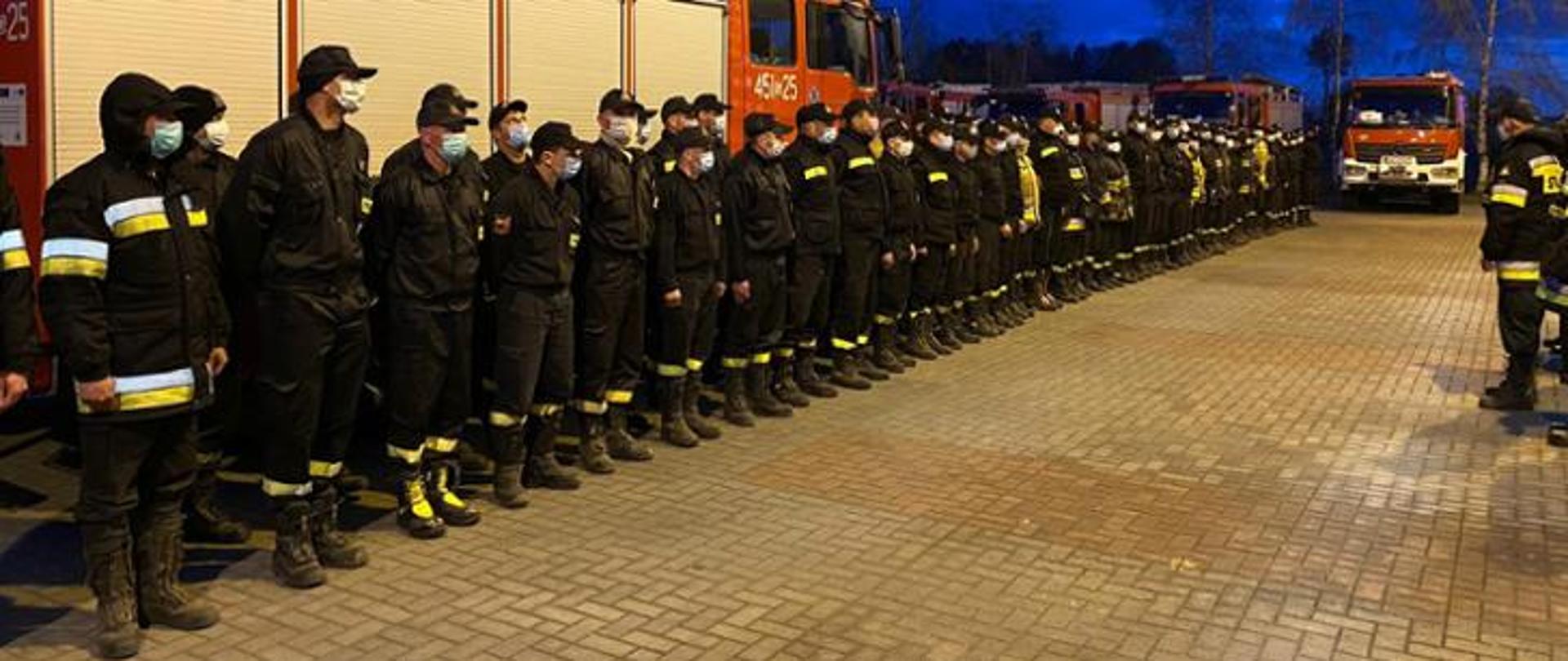 Na zdjęciu kilkudziesięciu strażaków ustawionych w dwuszeregu podczas zbiórki. Przed nimi stoją inni strażacy zwróceni twarzą do ustawionych w dwuszeregu. W tle pojazdy pożarnicze.