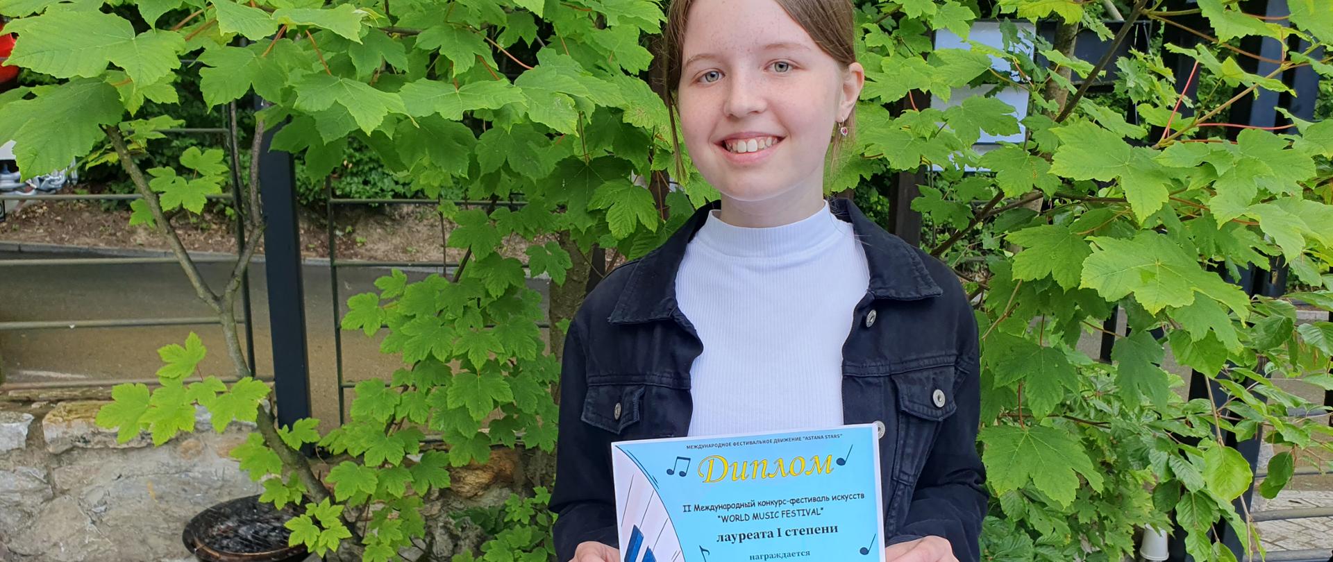 Uśmiechnięta uczennica w białym golfie ciemnej lekkiej bluzie trzyma w dłoniach dyplom w kolorze niebieskim z żółtymi napisami informujący o przyznaniu tytułu laureata I stopnia międzynarodowym konkursie online w Astanie. W tle drzewo klonowe zielone liście.
