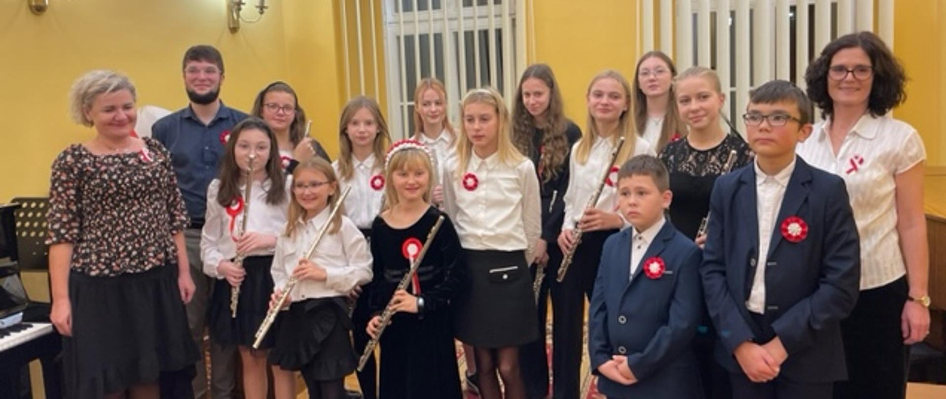 Grupa trojga nauczycieli oraz trzynaściorga uczniów. Część dziewczynek trzyma w dłoniach flety.