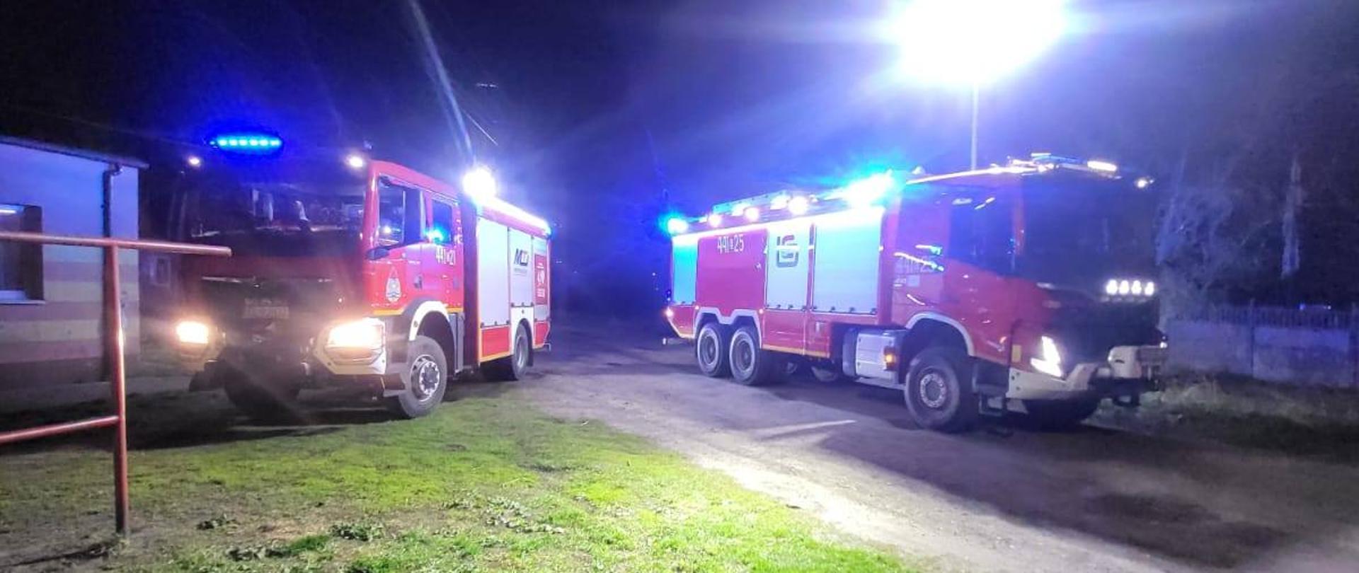 Na zdjęciu widoczne są dwa wozy strażackie. Jeden z nich stoi na drodze, drugi na trawie. Wozy mają włączone światła, dzięki czemu oświetlają okolicę.
