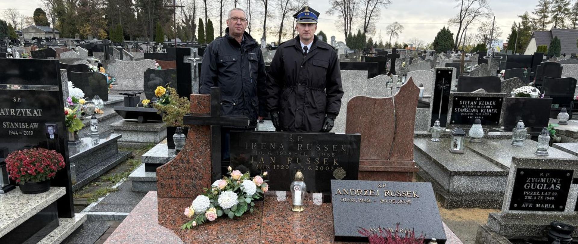 Na zdjęciu dwóch mężczyzn stoi przy nagrobku z czerwonego granitu na cmentarzu. Jeden z nich ubrany jest w mundur strażacki, drugi w ciemną kurtkę. Na grobie znajdują się znicze i kwiaty, a w tle widoczne są liczne inne nagrobki oraz drzewa bez liści.