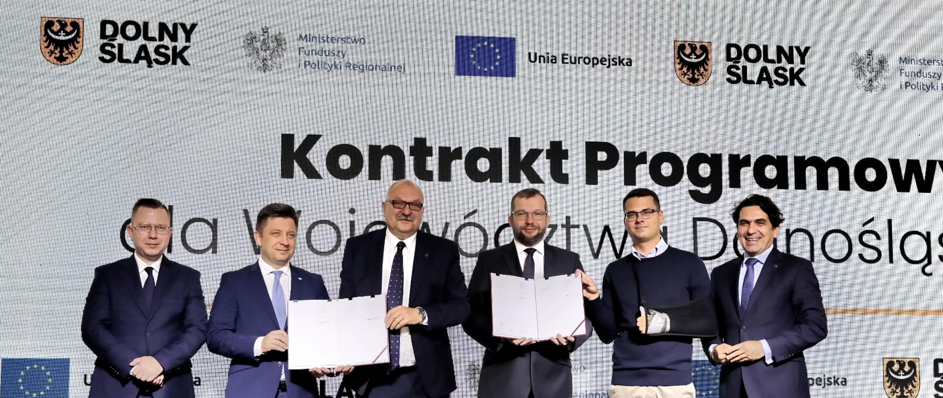  Zdjęcie przedstawia sześciu mężczyzn stojących obok siebie na scenie. Dwóch z nich trzyma w rękach podpisane dokumenty w twardej oprawie. Za ich plecami napis "Kontrakt Programowy". 