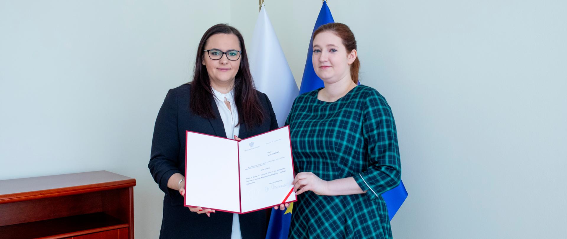 minister Małgorzata Jarosińska-Jedynak i wiceminister Anna Gembicka trzymają nominację, za nimi flagi Polski i Unii Europejskiej