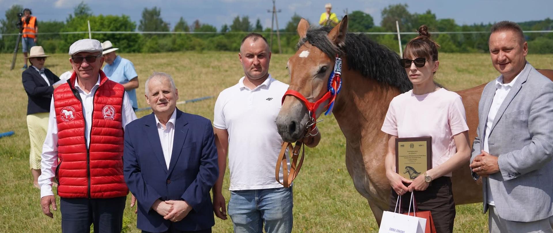 Kilka osób stoi przy koniu, jedna osoba trzyma w ręku dyplom i torby 