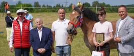 Kilka osób stoi przy koniu, jedna osoba trzyma w ręku dyplom i torby 