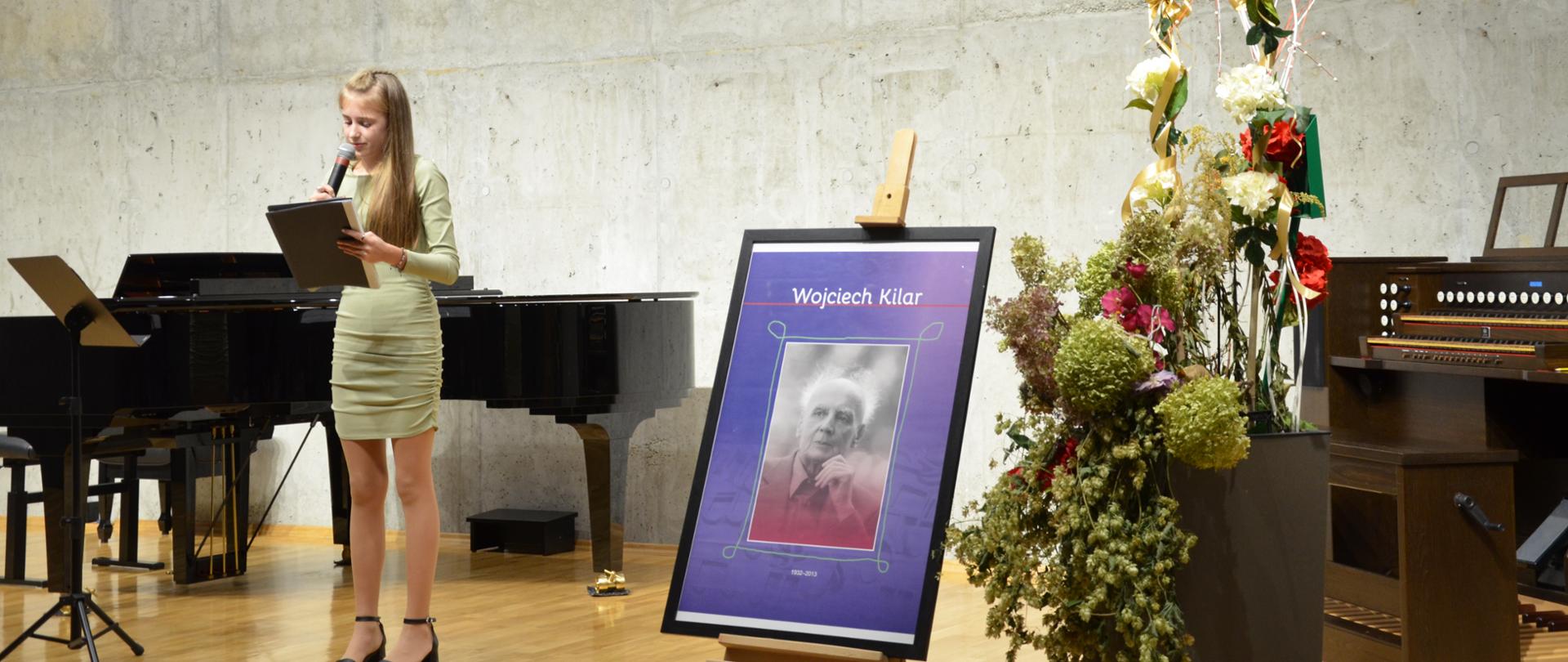 Uczennica stoi na scenie z mikrofonem, na pierwszym planie kosz z kwiatami, zdjęcie Wojciecha Kilara w biało-czerwonych barwach, na scenie organy i fortepian