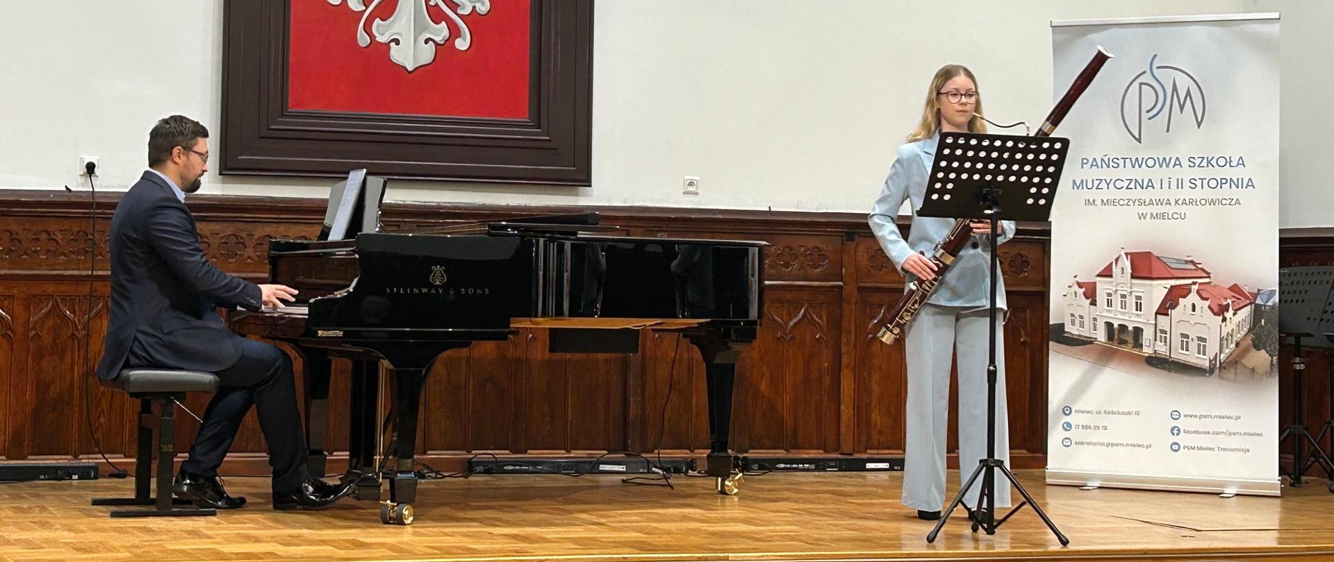 Uczennica gra na fagocie przy pulpicie nutowym, a nauczyciel akompaniuje na fortepianie Steinway podczas recitalu dyplomowego w Sali Królewskiej PSM w Mielcu.