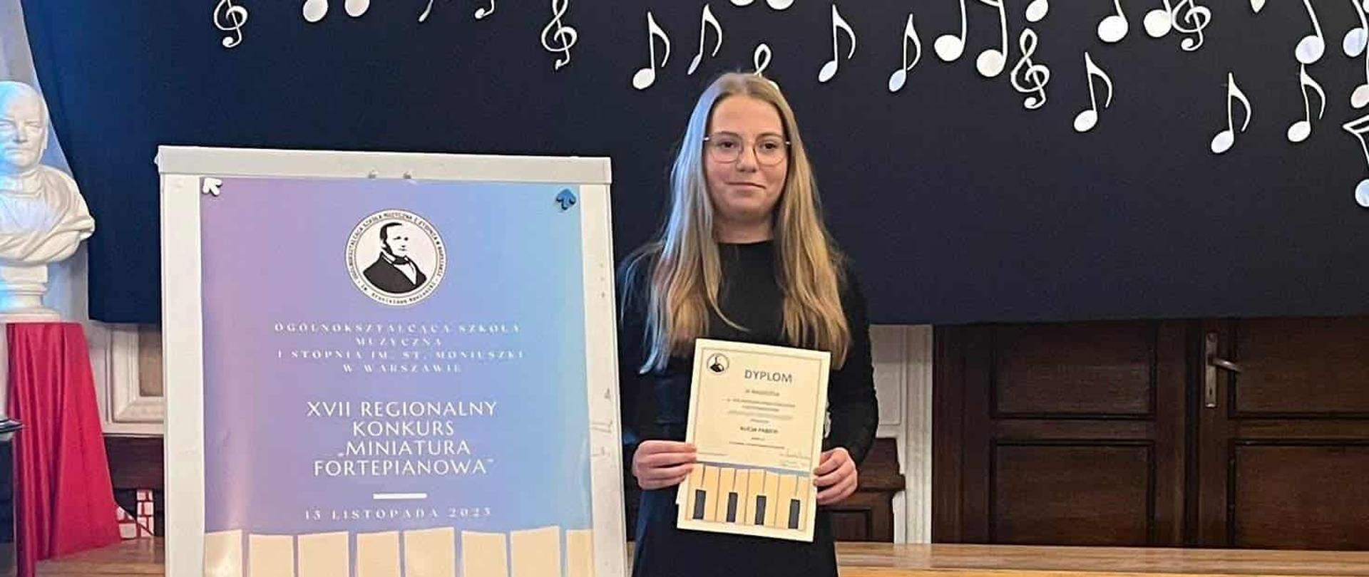 Uczennica z dyplomem po konkursie stoi na tle ściany w kolorowe nuty obok baner z nazwą szkoły muzycznej w której odbywał się konkurs