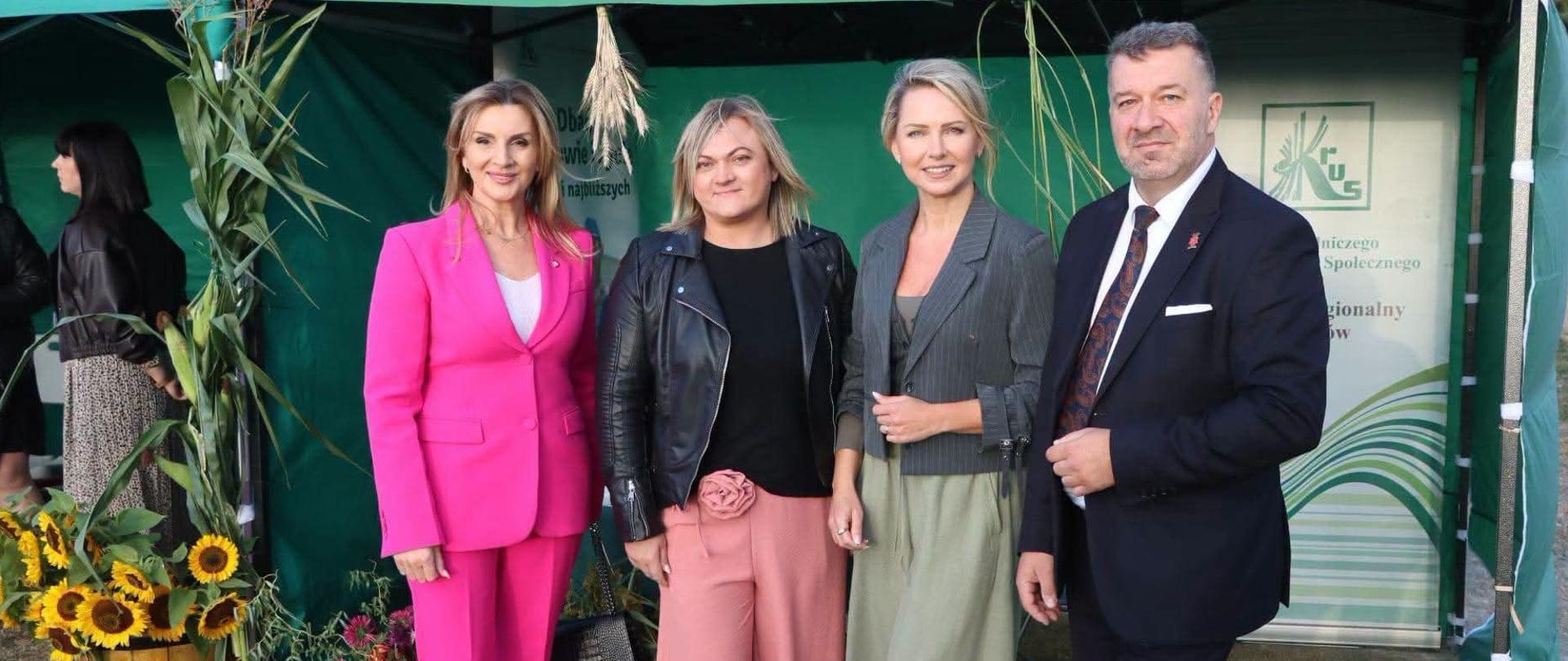 Trzy kobiety i mężczyzna pozują do zdjęcia przed stoiskiem Kasy pod zielonym namiotem z logo KRUS. Stoisko jest udekorowane żywymi kwiatami.