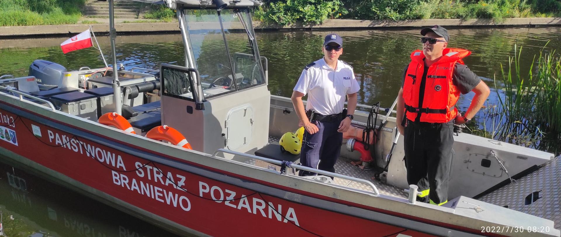 Rzeka. Na dużej łodzi strażackiej stoi strażak w pomarańczowej kamizelce i policjant w białej koszulce.