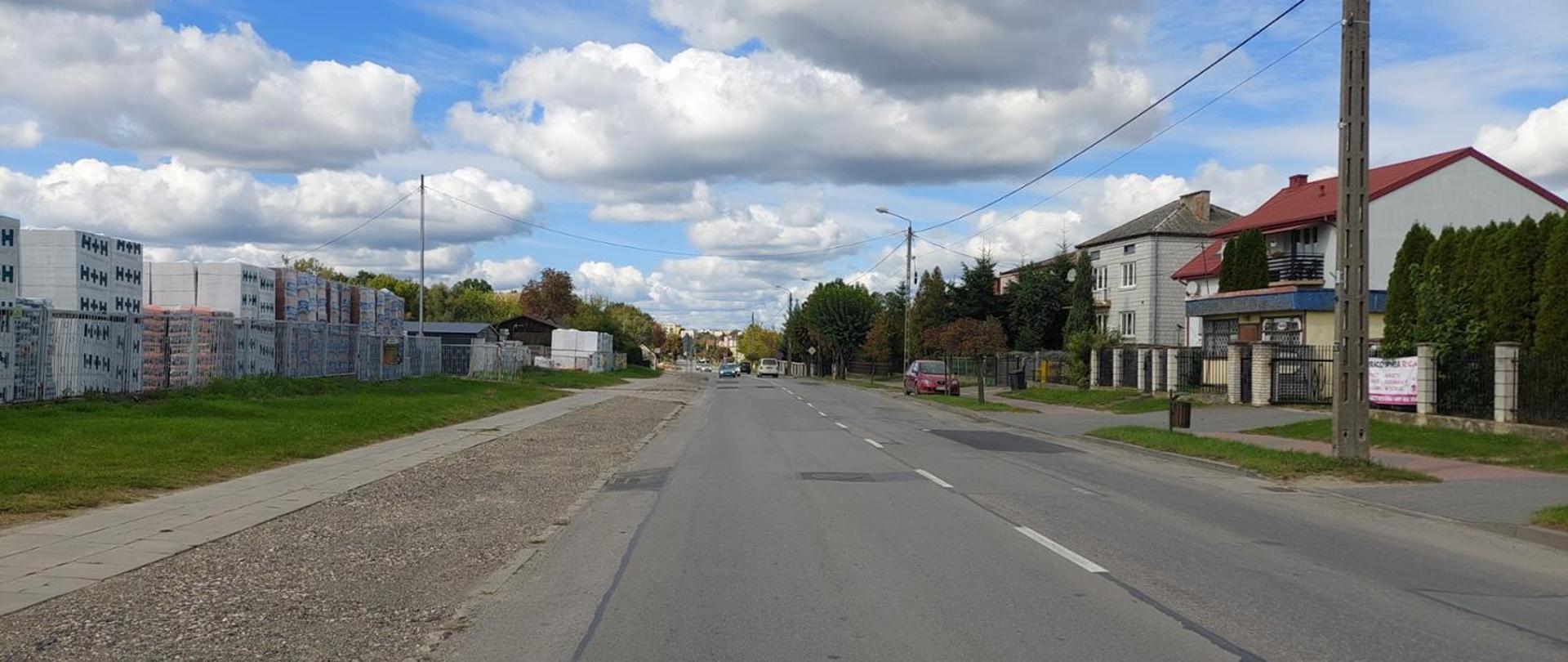 Widok z poziomu ziemi na drogę biegnącą obok niskich zabudowań. Ciepły, wiosenny dzień. Na niebie chmury.