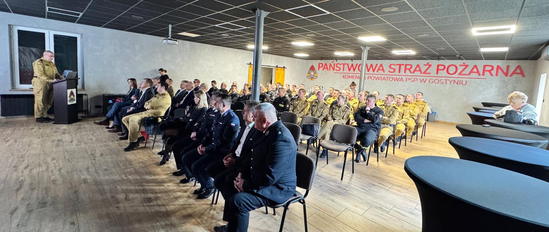 Komendant przemawia podczas odprawy. Zaproszone osoby siedzą w sali szkoleniowej.