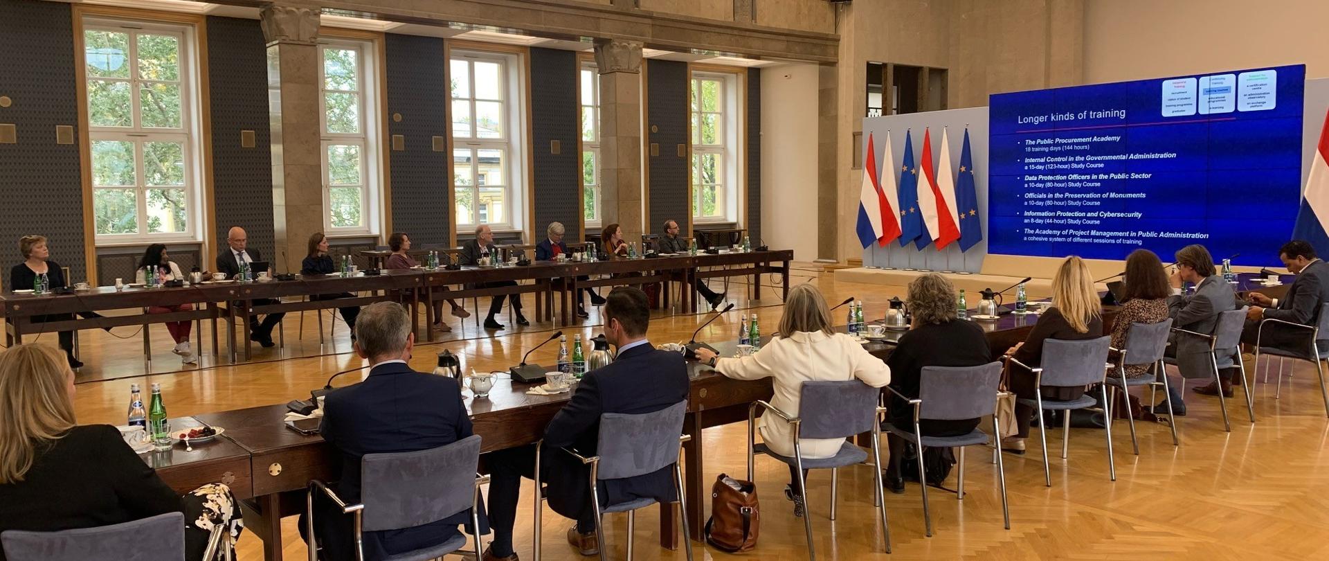 Na zdjęciu w wysokiej, eleganckiej sali przy stołach ułożonych w półokrąg siedzi kilkanaście osób, elegancko ubranych. Na stołach widać mikrofony, butelki z wodą. Za uczestnikami spotkania znajdują się wysokie okna. Na środku sali jest duży telebim z wyświetlaną prezentacją, po jego lewej stronie flagi Polski, Królestwa Niderlandów oraz Unii Europejskiej.