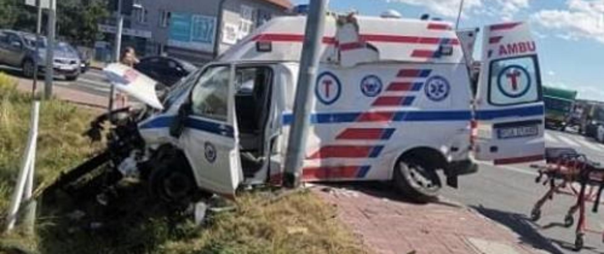Karetka po wypadku częściowo znajduje się w rowie oraz na chodniku. Widoczne duże zniszczenia karoserii pojazdu.