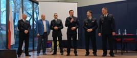 Funkcjonariusze Państwowej Straży Pożarnej w mundurach wyjściowych stoją obok siebie z mężczyzną w garniturze za nimi ekran do wyświetlania prezentacji oraz stół na którym są butelki z wodą.