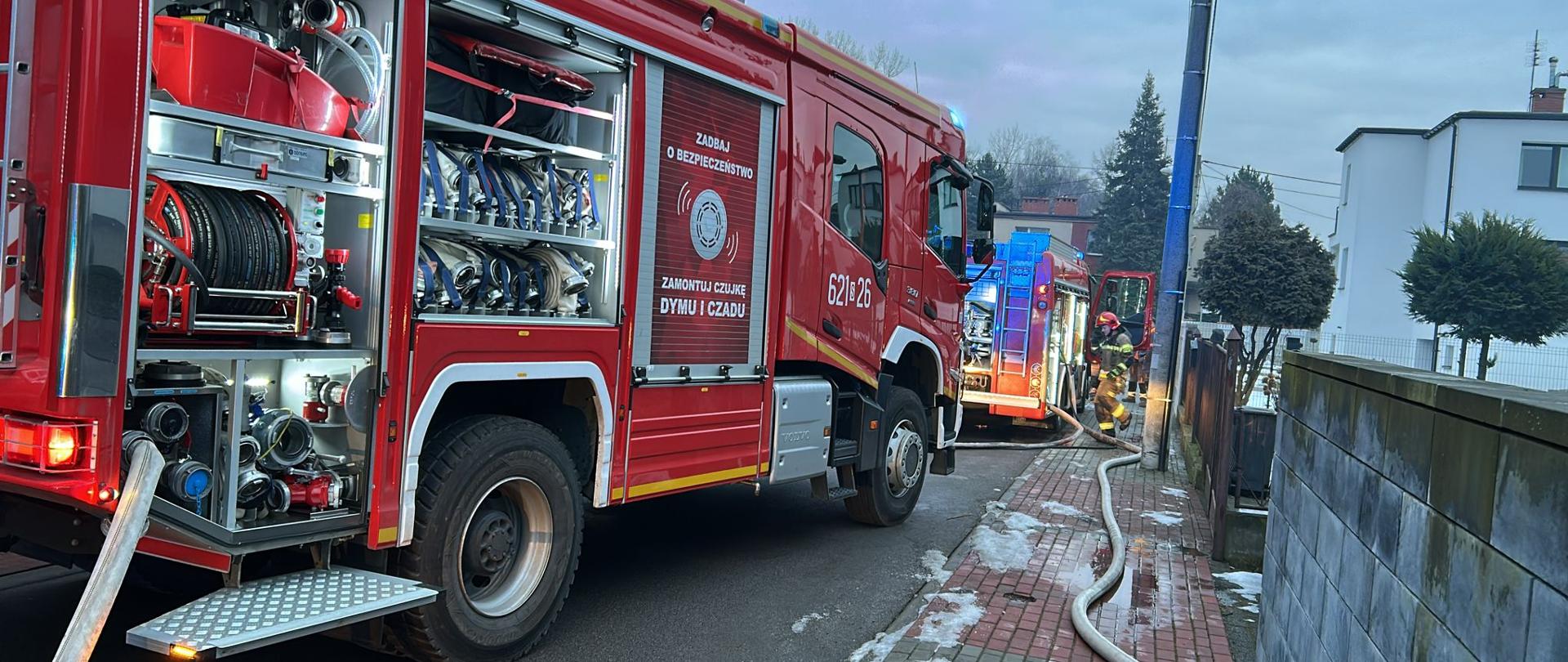 Zdjęcie przedstawia akcję służb ratunkowych na osiedlowej ulicy w zimowej scenerii. Na pierwszym planie dominuje duży, czerwony wóz strażacki z otwartymi skrytkami bocznymi, w których widać profesjonalny sprzęt: węże tłoczne, zwinięte linie szybkiego natarcia oraz specjalistyczne oprzyrządowanie pompowe. Na boku pojazdu widnieje numer operacyjny oraz plakat edukacyjny zachęcający do montażu czujek dymu i czadu, co podkreśla prewencyjny i ratowniczy charakter misji.
W głębi kadru, na mokrym od roztopionego śniegu chodniku, widać rozciągnięte węże strażackie prowadzące w stronę posesji. Drugi wóz strażacki stoi kawałek dalej z włączonymi niebieskimi sygnałami błyskowymi, a obok niego pracuje strażak w pełnym umundurowaniu bojowym i hełmie. Całość sceny, osadzona w chłodnym, popołudniowym świetle przy niskiej zabudowie mieszkalnej, oddaje powagę sytuacji i dynamikę działań straży pożarnej.
