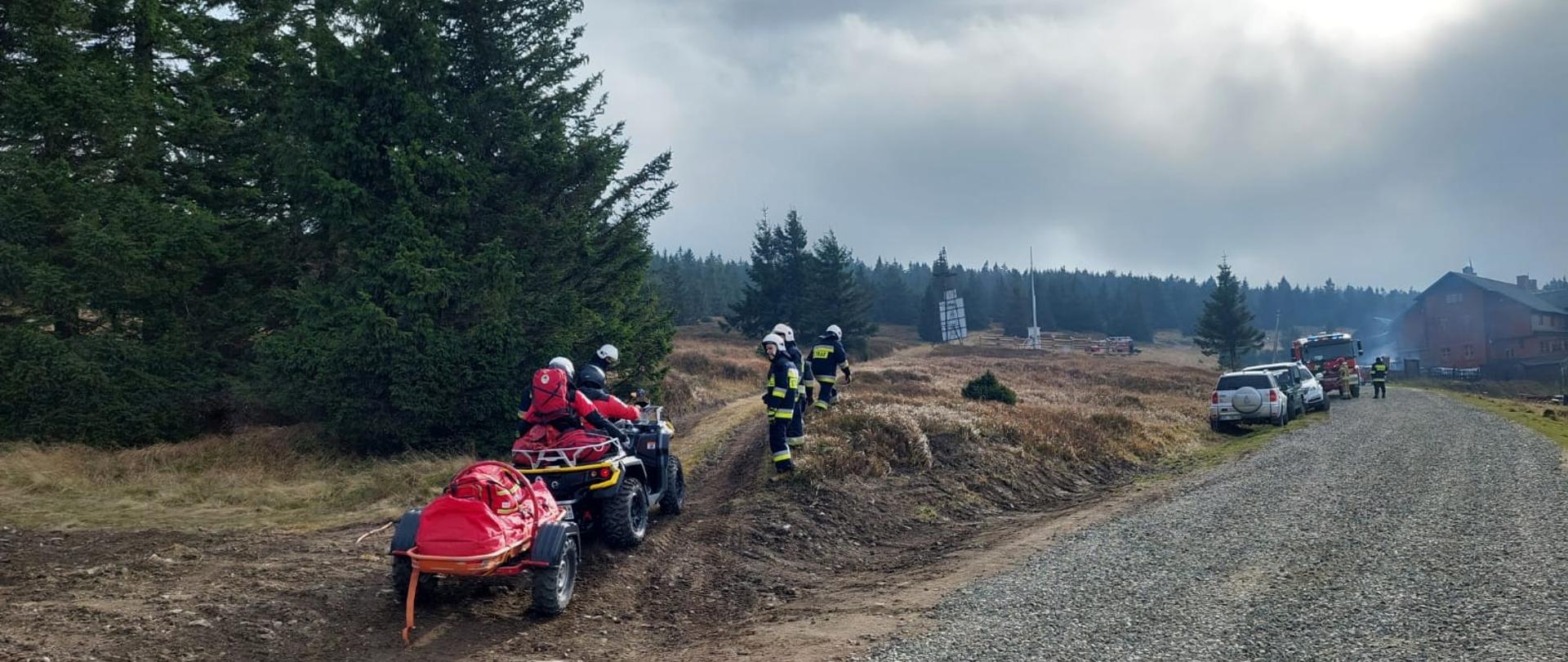 Po prawej stronie planu biegnie kamienista droga. Po lewej stronie drogi po nasypie na quadzie z przyczepką ratowniczą poruszają się ratownicy. W oddali po lewej stronie drogi stoją zaparkowane dwa samochody osobowe i samochód pożarniczy.