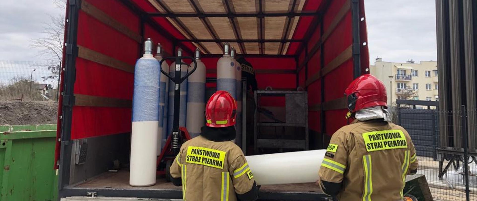 Zdjęcie przedstawia dwóch strażaków wyładowujących butle z tlenem z samochodu dostawczego. Pod plandekom za nimi widać kolejne butle z tlenem.