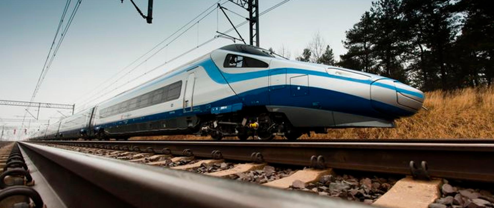 Pociąg Pendolino pędzący po torach. Na dole widać tory kolejowe. Na środku pociąg. W tle rozciągający się po horyzont szlak kolejowy.