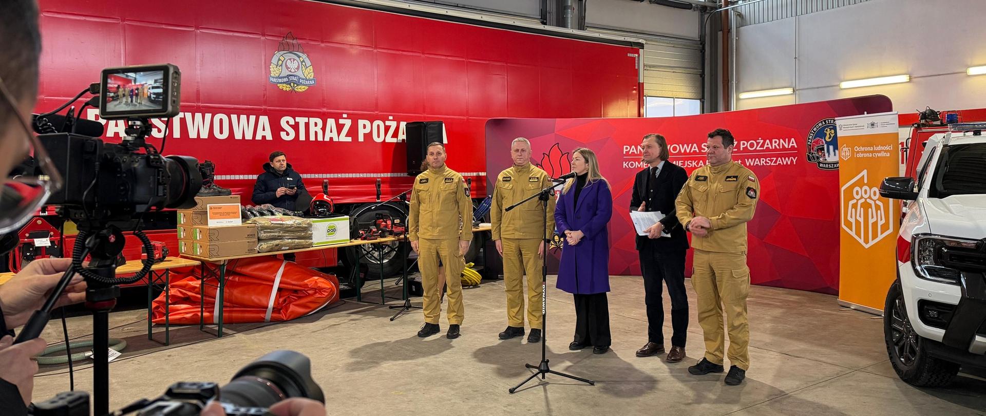 Szerokie ujęcie wnętrza hangaru straży pożarnej. Na środku stoi grupa pięciu osób: trzech strażaków w piaskowych mundurach służbowych, kobieta w fioletowym płaszczu oraz mężczyzna w ciemnym garniturze trzymający teczkę. Przed nimi ustawiony jest mikrofon na statywie. W tle widać wielką czerwoną naczepę z napisem „PAŃSTWOWA STRAŻ POŻARNA”, stół ze sprzętem ratowniczym oraz biały samochód terenowy. Na pierwszym planie widoczne są plecy i ręce operatora kamery filmującego wydarzenie.