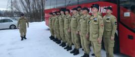 Strażacy stoją w dwóch rzędach przed autobusem niedaleko nich stoi dowódca grupy.