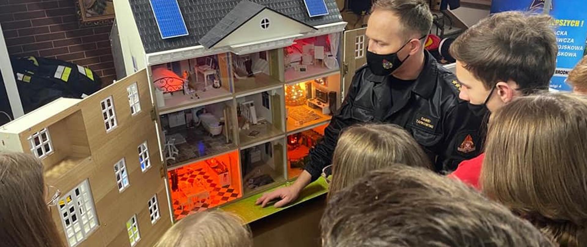 GRUPA DZIECI SKUPIONYCH PRZY OSOBIE STRAŻAKA W UBRANIU DOWÓDCZO-SZTABOWYM, STOJĄCEGO PRZY MOBILNYM SYMULATORZEZ ZAGROŻEŃ POŻAROWYCH