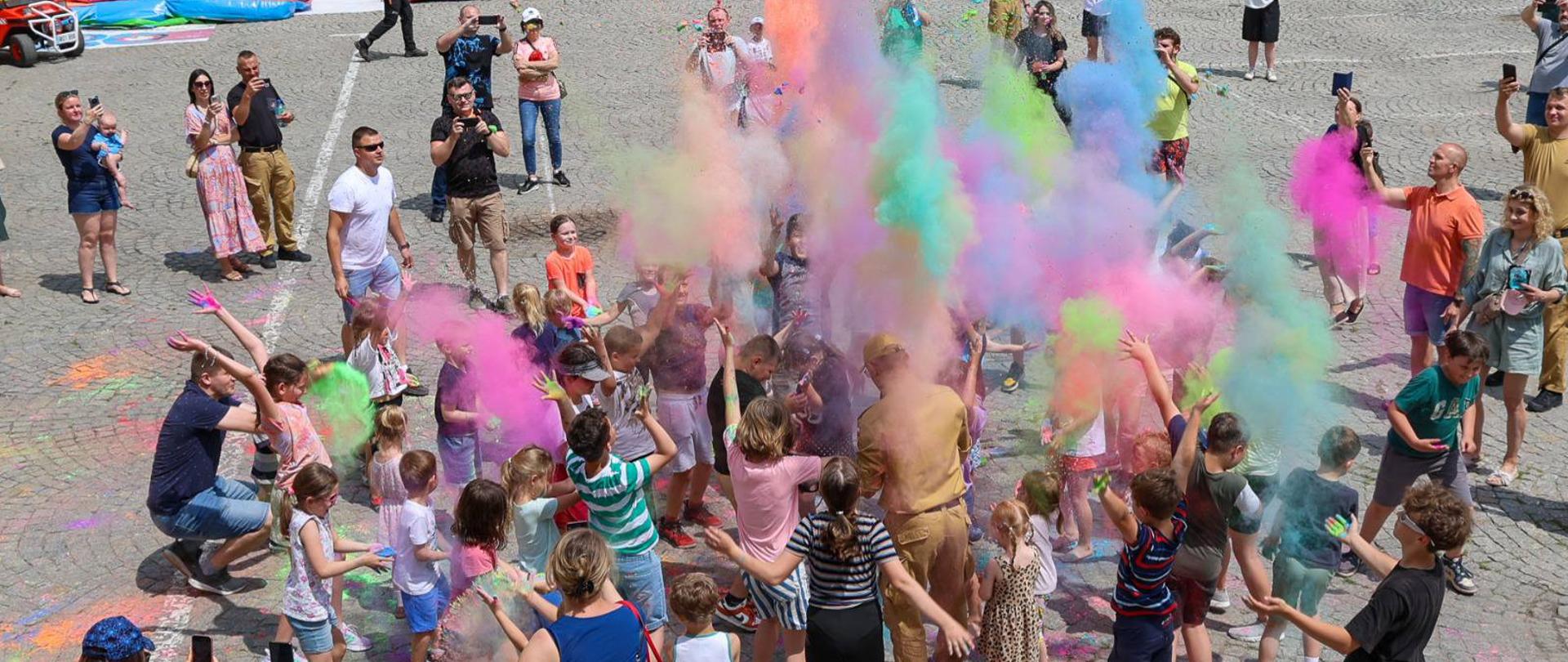 Na placu przed budynkiem szkoły dzieci i dorośli uczestniczą w zabawie z okazji Dnia Dziecka. Na środku placu widać grupę osób którzy wyrzucają w górę kolorowy proszek – festiwal kolorów. 