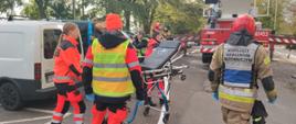 Ratownicy medyczni z pustymi noszami idą do poszkodowanego, obok po prawej znajduje się strażak w kamizelce kierujący działaniami ratowniczymi, w tle stoi podnośnik hydrauliczny.
