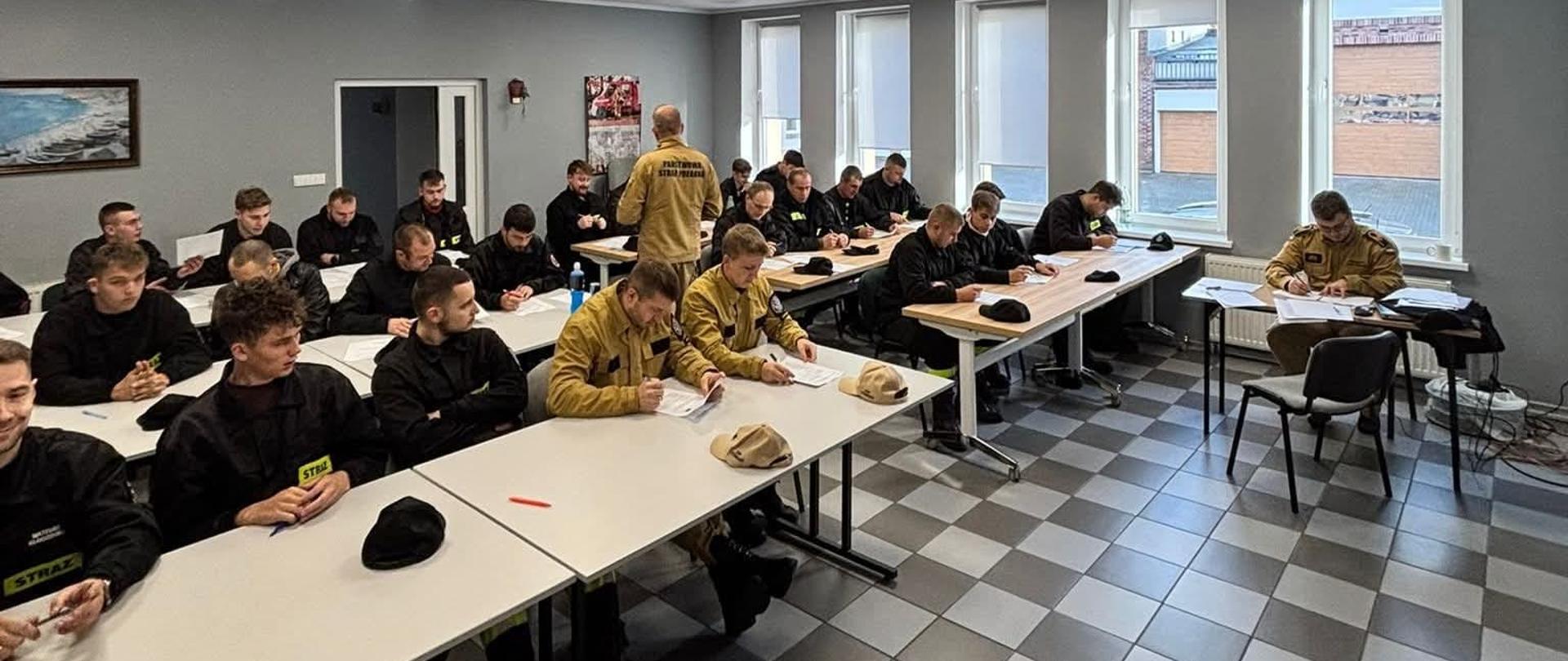 Druhny i Druhowie z jednostek OSP piszą egzamin kończący szkolenie ratowników OSP. Egzamin odbywa się w sali edukacyjnej OSP Puck. Strażacy są ubrani w stroje koszarowe w kolorze czarnym.