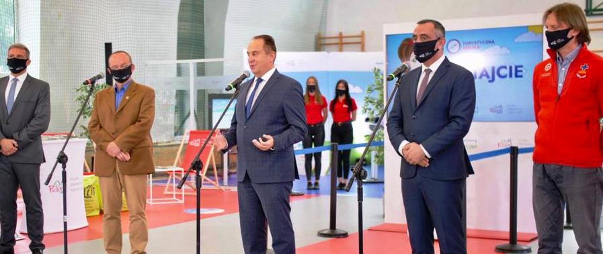 Wiceminister Andrzej Gut-Mostowy stoi w środku przed mikrofonem w sali gimnastycznej w szkole i opowiada o projekcie "Turystyczna Szkoła".