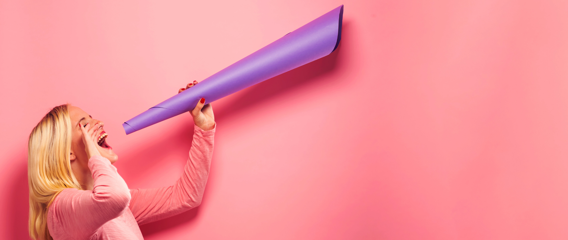 Z lewej strony odwrócona bokiem kobieta mówiąca do zrobionej z fioletowego papieru tuby. Tło zdjęcia także różowe.