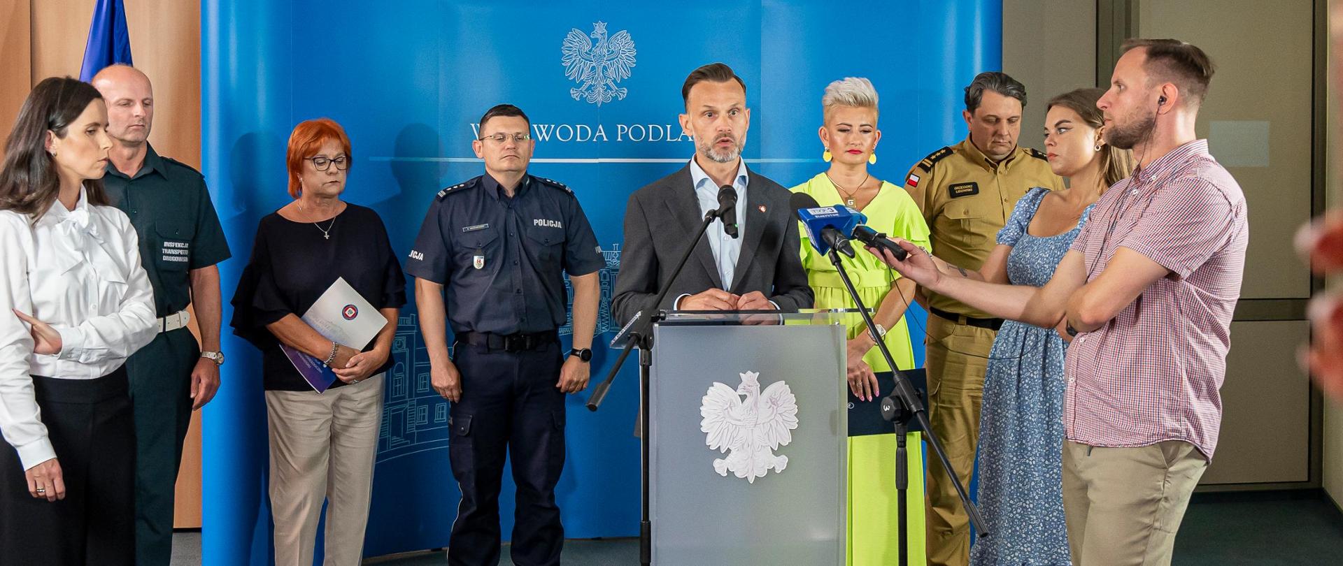Konferencja prasowa "Bezpieczne wakacje" w Podlaskim Urzędzie Wojewódzkim. Przy mównicy z godłem wojewoda podlaski Jacek Brzozowski. Za im na tle niebieskiej ścianki pozostali uczestnicy konferencji.