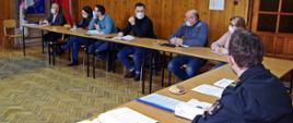 Widok z przodu - na sali szkoleniowej przy stolikach siedzą uczestnicy spotkania. Widok od tyłu – przy stoliku siedzi zastępca komendanta powiatowego PSP.
