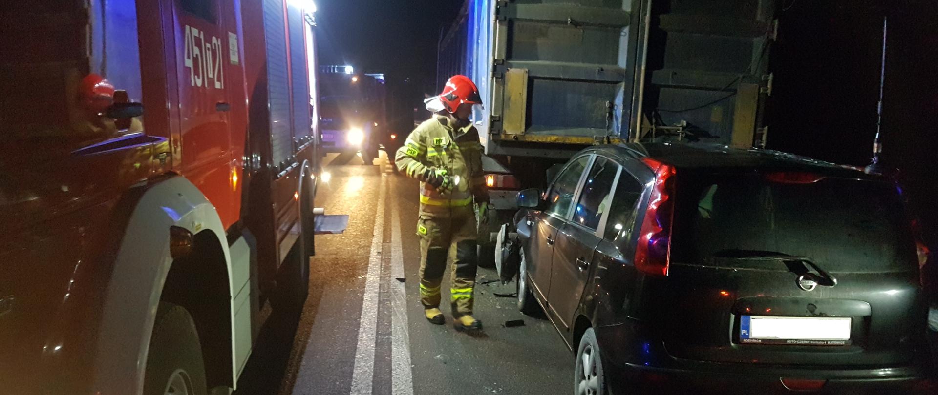Na zdjęciu w porze nocnej widać strażaka, który stoi obok samochodu osobowego, który wjechał w tył samochodu ciężarowego. Przód samochodu osobowego jest rozbity. Z lewej strony zdarzenia zaparkowane zostały samochody strażackie