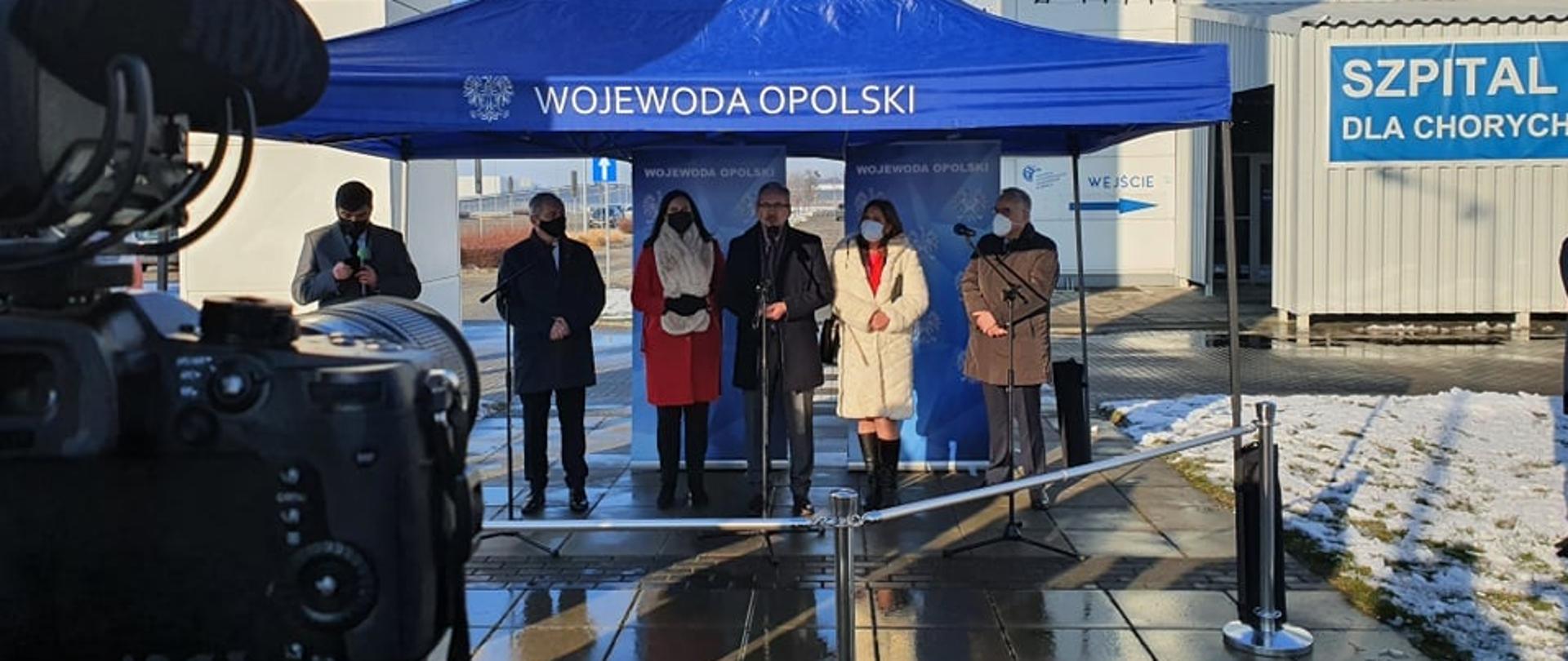 Konferencja prasowa ministra zdrowia. Na zdjęciu 6 osób, stojących pod granatowym namiotem z napisem Wojewoda Opolski. Przed nimi mikrofony dziennikarskie. Trwa konferencja prasowa. Na środku kadru stoi minister zdrowia, który udziela wypowiedzi. W tle budynek szpitala tymczasowego w Opolu. Po lewej stronie kadru z przodu widać stojącą na statywie kamerę telewizyjną.